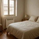 Dormitorio de invitados con una cama, un radiador eléctrico junto a la pared y una manta doblada sobre una silla.
